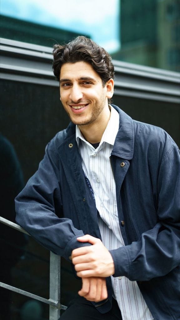 Example photo: Smiling man with curly dark hair in a navy jacket, leaning on a city railing