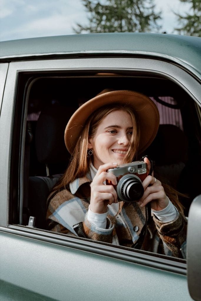 Example photo: Smiling woman in a brown hat holding a film camera at a car window