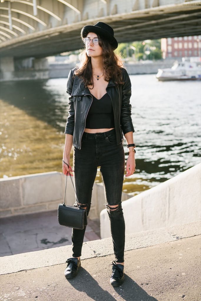 Example photo: Woman in a black hat, leather jacket and ripped jeans standing full body by a river under a bridge