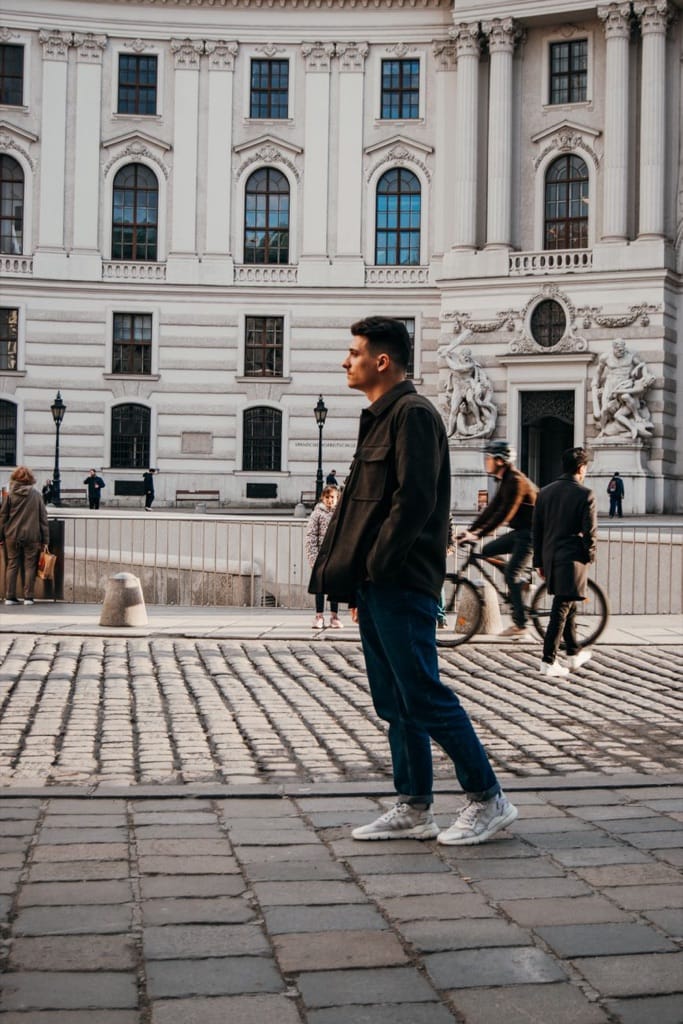 Example photo: Man in a dark jacket standing on a cobblestone square by a grand old building, passers-by behind him